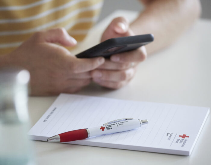 Zwei Hände halten ein Handy in der Hand. Im Vordergrund des Bildes sieht man einen Block und einen Kugelschreiber. Auf beidem ist das Logo vom DRK in Bonn aufgedruckt.
