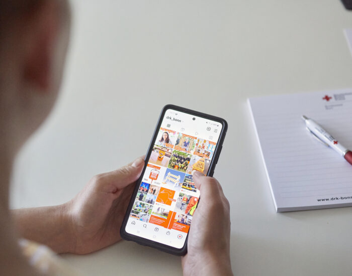 Zwei Hände halten ein Handy in der Hand. Darauf erkennt man einen Ausschnitt vom Instagram-Feed vom DRK in Bonn.