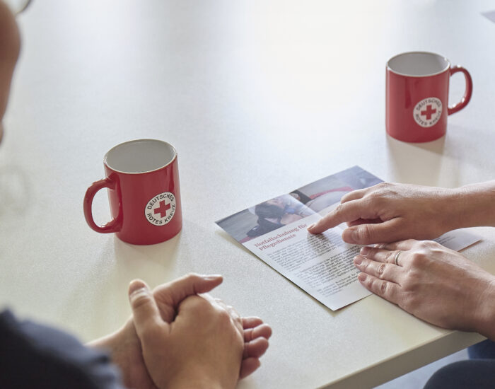 Die Hände von zwei Personen sind zu sehen, ebenfalls ein Flyer. Eine der Hand weist auf einen Text in dem Flyer. Im Hintergrund stehen zwei rote Tassen auf einem Tisch.