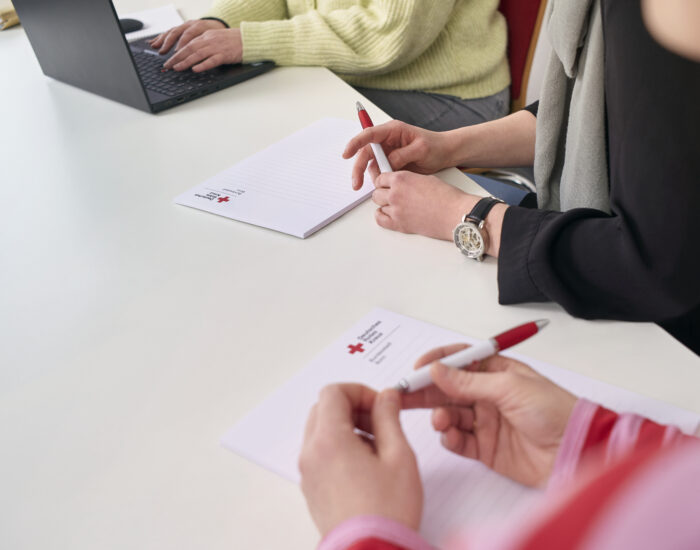Menschen sitzen am Tisch und haben Kugelschreiber in der Hand. Man sieht Blöcke und Hände, die die Stifte festhalten. Im Hintergrund ist ein Laptop.