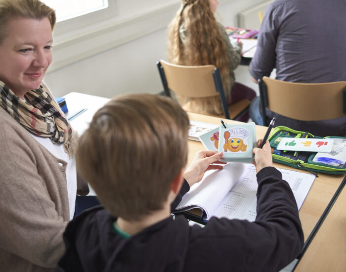 Ein Junge sitzt am Schultisch und arbeitet mit Hilfekarten. Eine Schulbegleiterin sitzt neben ihm.