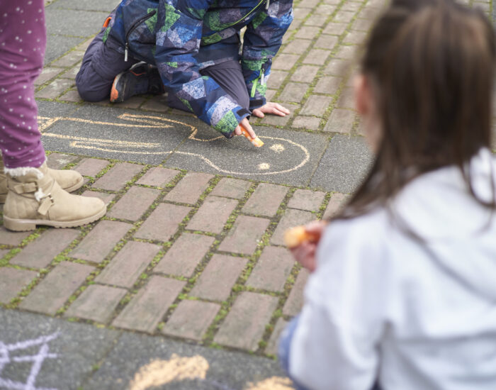 Drei Kinder, von denen eines mit Kreide auf den Boden malt.