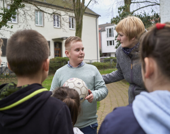 Ein Kind mit einem Fußball in der Hand steht, gemeinsam mit einer Schulbegleiterin, vor einer Gruppe Kinder.