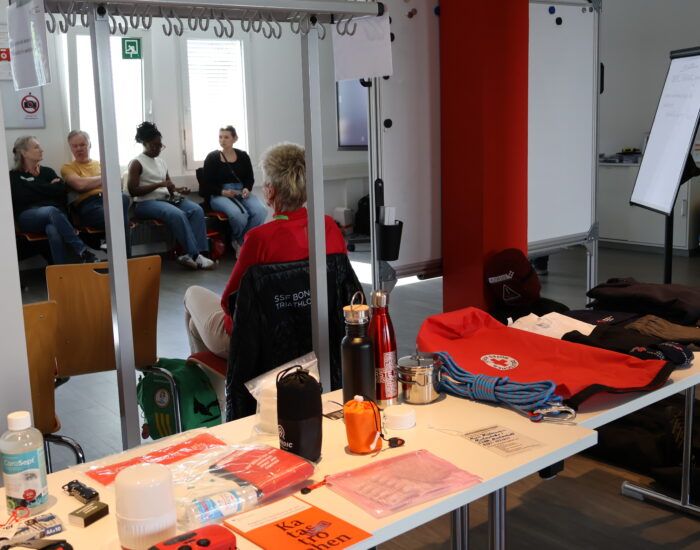 Im Vordergrund ein Tisch mit Utensilien eines Notfallrucksacks, im Hintergrund Personen auf Stühlen.